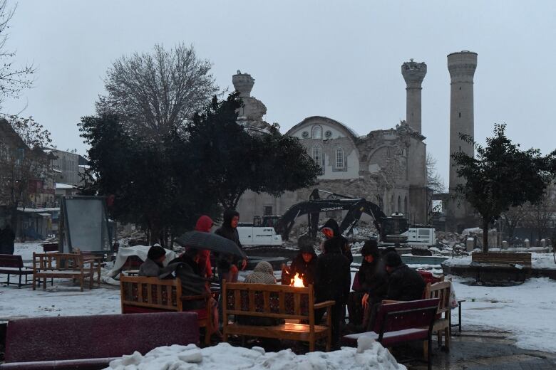 People sit on benches outdoors, surrounding a small fire, with a damaged concrete structure in the background.