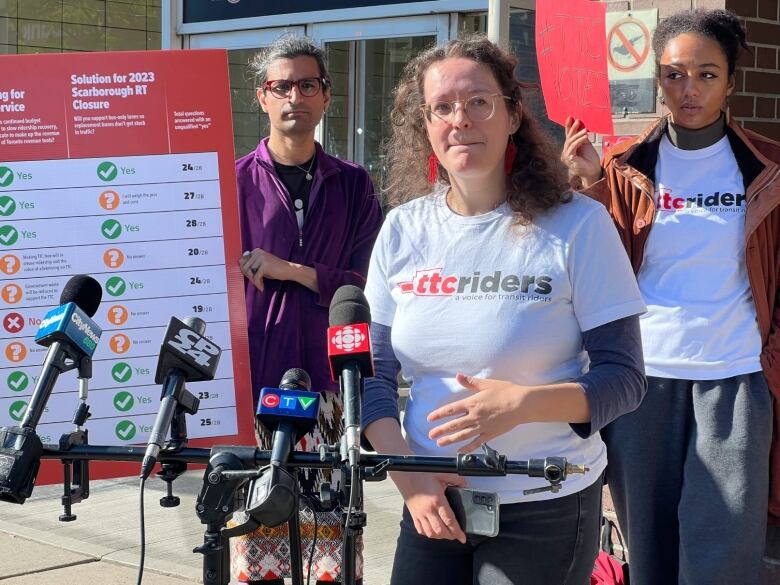 A woman with long, curly hair wearing a shirt that reads TTCriders, stands in front of a bank of microphones with two people in similar shirts holding signs. 