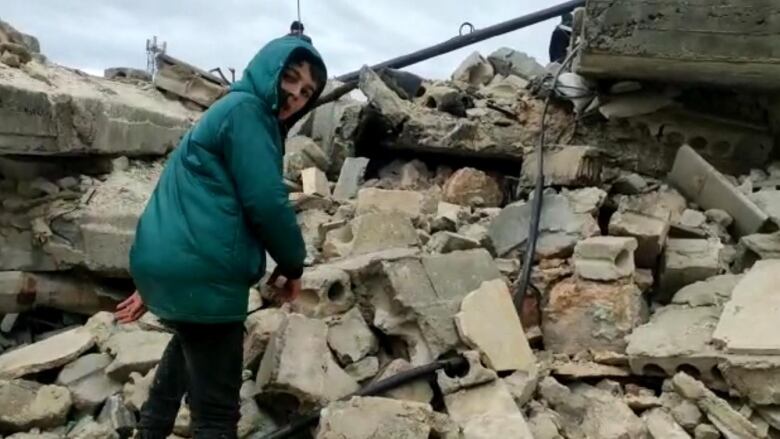 A young boy in a bright blue parka peers over his shoulder as he climbs a mound of rubble. 