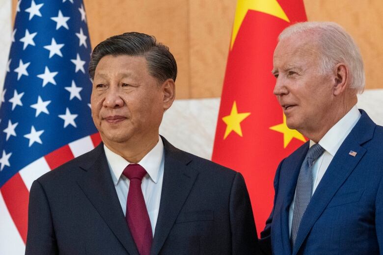 A man wearing a black suit and a burgundy tie stands next to a man wearing a blue suit and tie, with American and Chinese flags in the background.