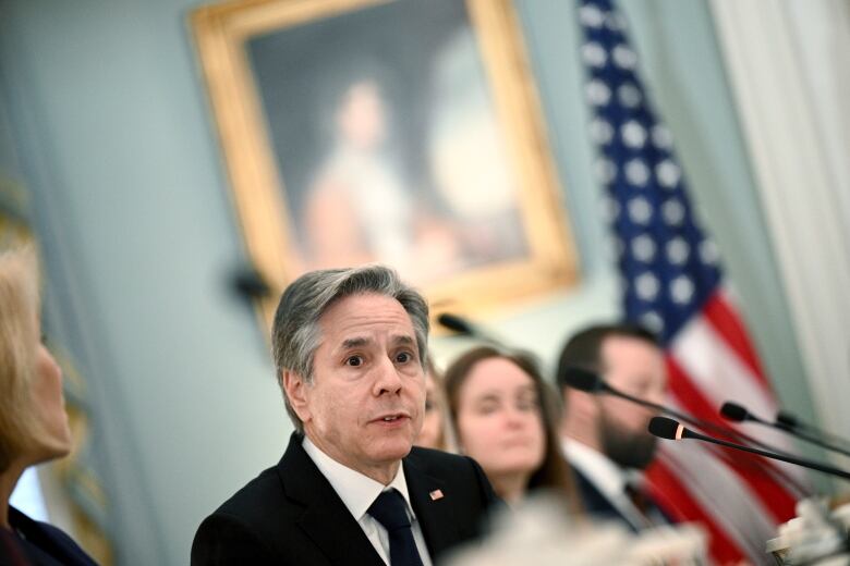 A man wearing a black suit and tie sits at a table, with others around him, in front of a small microphone. A painting and an American flag can be seen in the background.