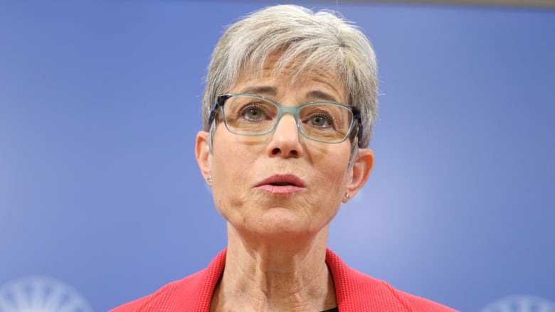 A woman with white hair wearing a red blazer covering a blue and white polka dot blouse speaks during a news conference.