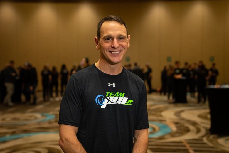 A man standing in a hotel ballroom in a Team P.E.I. shirt. 