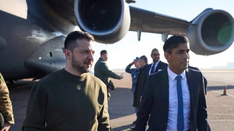 Two men are shown on a tarmac near a plane, one in a sweatshirt, the other in a suit.