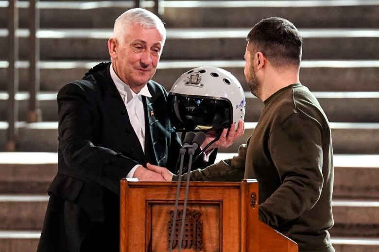 A man hands an older man a white helmet, near a lectern.