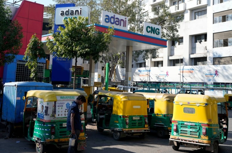 Four yellow and green rickshaws line up at a compressed natural gas station, with the name 'Adani' written on a white, red and blue overhang. A white apartment building is shown in the background.