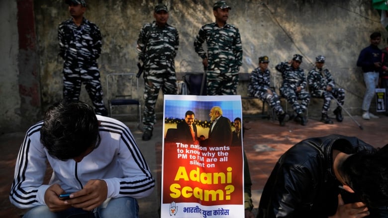 Two men sit on a curb, as a poster bearing the image of two men in suits is shown between them, with six people behind them, dressed in black and white fatigues. The posted reads: 'The national wants to know why the PM to FM are all silent on Adani scam?'