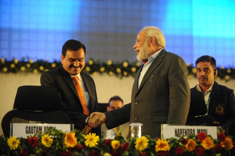 Two men wearing suits shake hands, as a security guard looks on behind them.