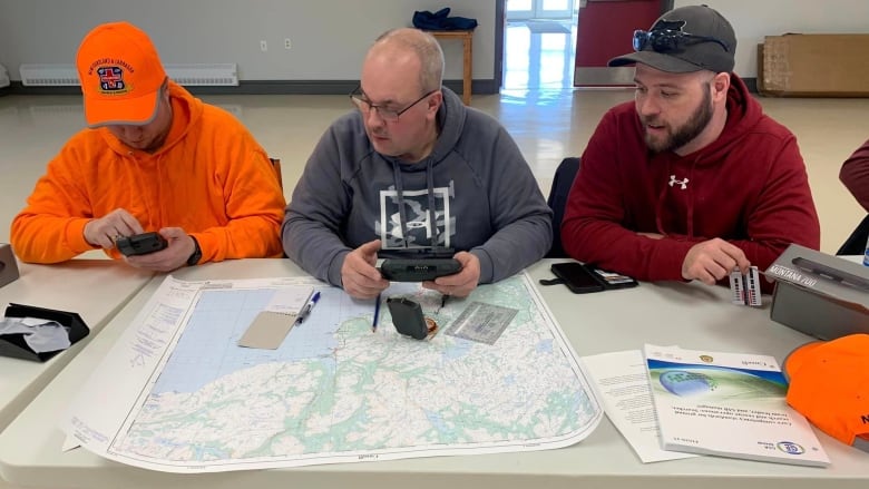 Three men, one dressed in neon orange, sit at a table examining maps and a compass.