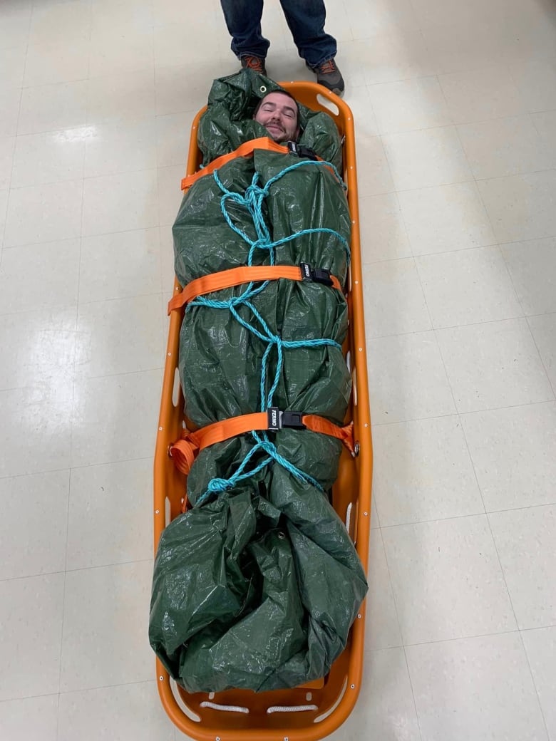 A man smiles while lying bundled up in a tarp in a search and rescue stretcher.