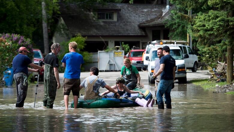 Three people sit in a raft floating in about 30 cm of floodwater in centre frame with five more people gathered around.