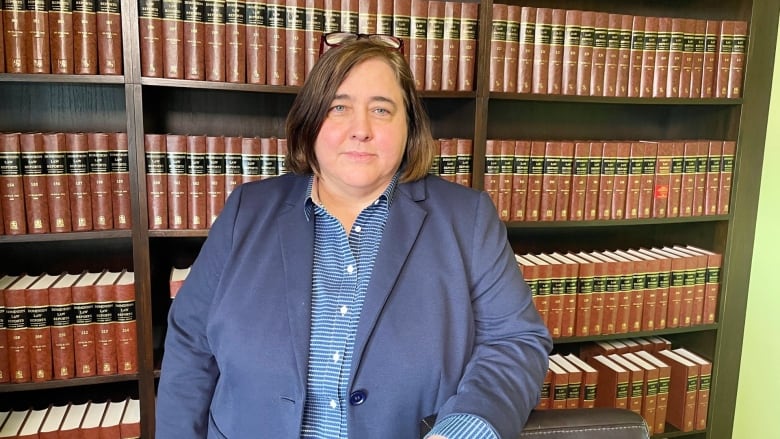 A white woman wearing a blue patterned collar shirt under a blue blazer sits in front of a bookshelf filled with black and brown-spined books. Her facial expression is neutral, and she has mid-length brown hair with a pair of glasses perched on top of her head.