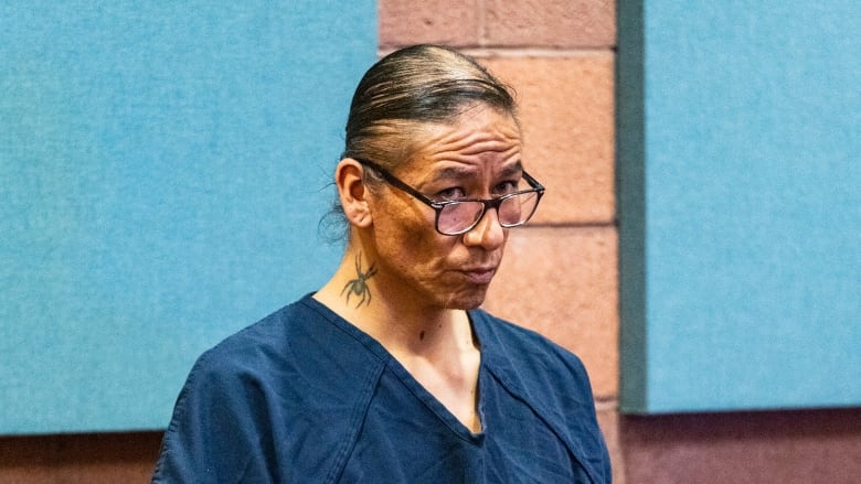 A man in black glasses and a dark blue prison jumpsuit stands in court.