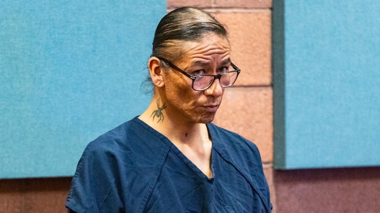 A man in black glasses and a dark blue prison jumpsuit stands in court.