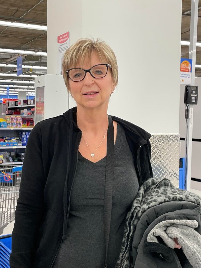 A blonde woman with a pixie cut dressed in gray and black standing inside a grocery store. 