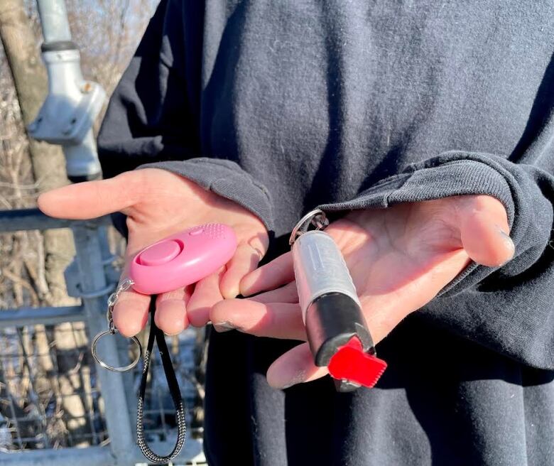 A woman holds a small pink alarm on a keychain and a tube of coyote spray.