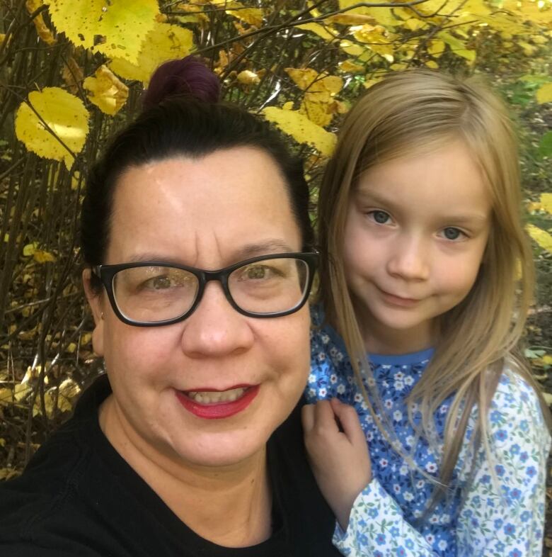 A middle-aged woman with glasses smiles next to her young, blonde-haired daughter while tree branches in the background bear fall colours. 