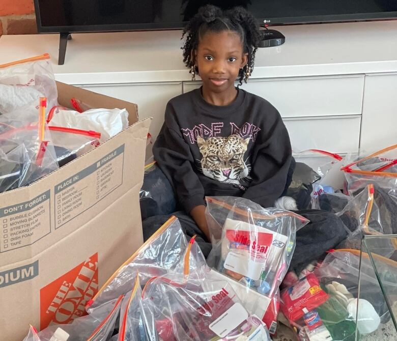 A young girl sits surrounded by bags stuffed with water bottles, snacks, gloves and other winter essentials.