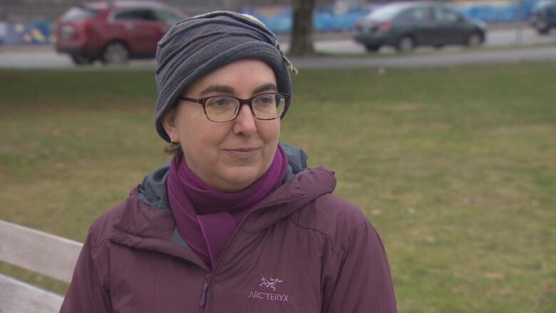 A woman in a grey hat wearing a purple jacket sits on a park bench during an interview.