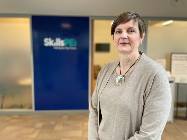 Woman with short brown hair wearing beige sweater set stands in front of blue banner that says Skills P.E.I.