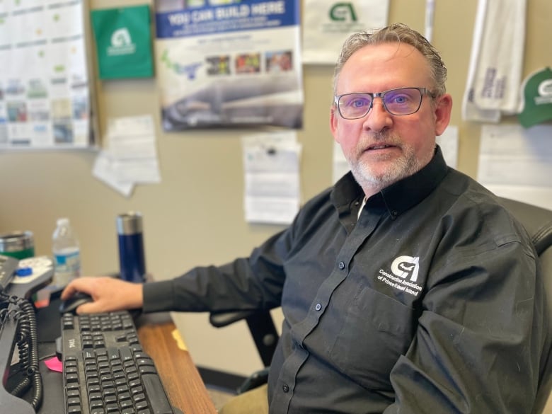 Man with short grey hair and glasses sits at computer wearing a dark shirt with branded logo for the Construction Association of P.E.I.