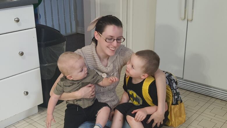 A mom wearing glasses is shown sitting in a kitchen with her two sons. One of them on her left knee is wearing a batman t-shirt.