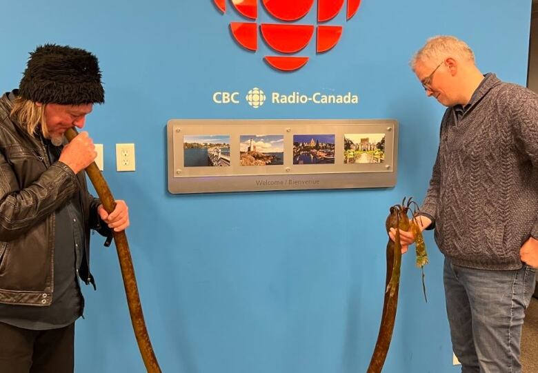 Two men hold the ends of a long piece of kelp, while one blows into it