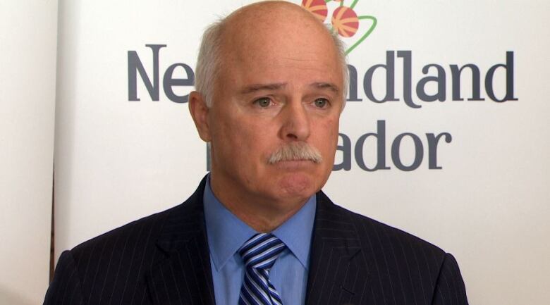 A man wearing a black suit jacket and a blue shirt standing in front of a banner which reads Newfoundland and Labrador. 