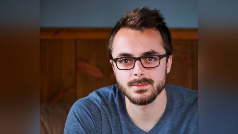 A man with glasses and goatee in a blue pullover stands in front of a wooden wall.