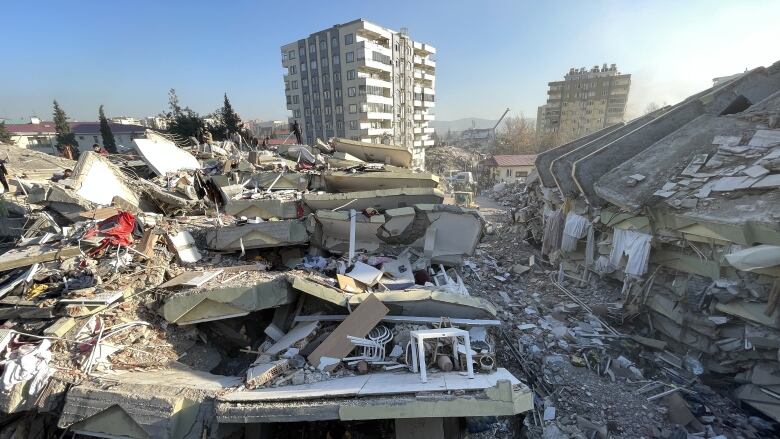 The wreckage of flattened buildings are shown, including plastic chairs and tables, as well as fabric bits, while a white apartment building still stand further back, against a blue sky.