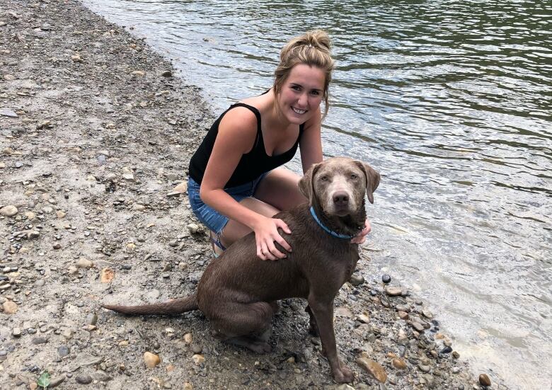 a woman squats next to a dog near a body of water