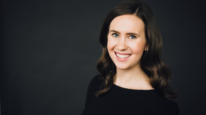 a woman with brown hair smiles. she has a black shirt and is in front of a black background 