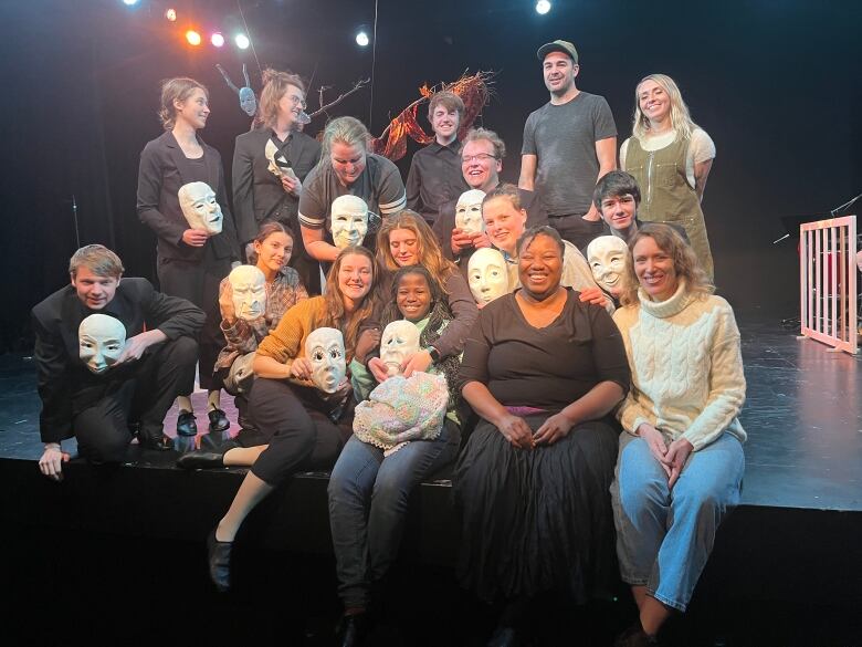 The cast sit on the edge of the stage. One girl has her arms wrapped around Vold, as Vold smiles widely toward the camera. Many of the cast are holding white masks, a metaphor used in the play to show how Vold masks her emotions to protect herself and her loved ones. 