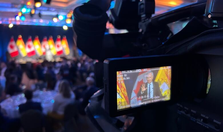 A camera with a man on the viewfinder, with a blurry stage with flags on it in the background.