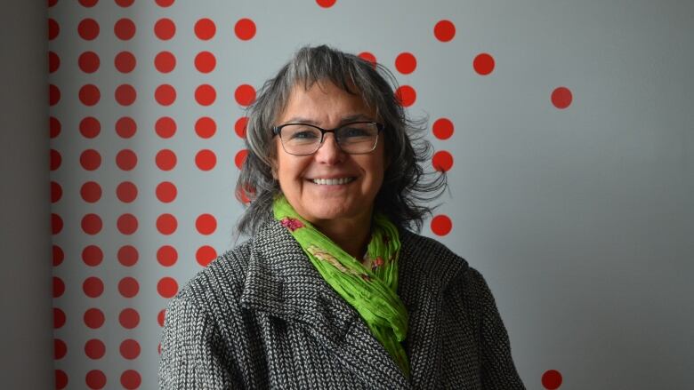 A woman wearing a grey coat and a green scarf smiles into the camera.