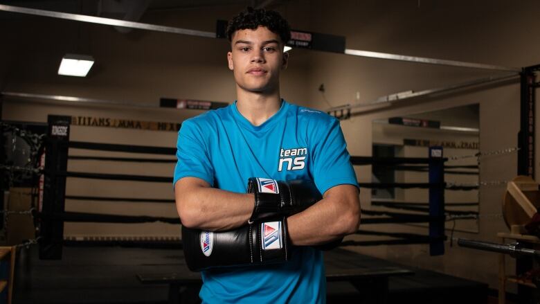 A teenager in a blue Team Nova Scotia T-shirt stands outside a boxing ring with his arms crossed.