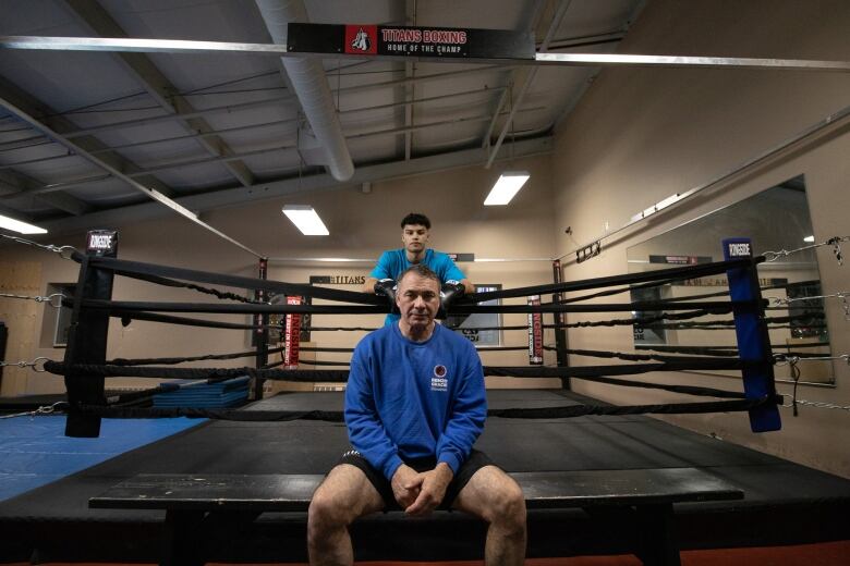 An boxing coach sits outside the ring, while a boxer he coaches stands behind him. 