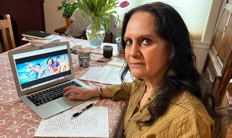 A woman sits at a table, staring at the camera. On the table in front of her is a laptop, with an image from a film on screen. 