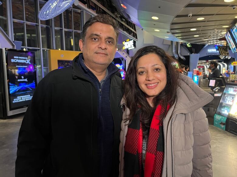 A man and a woman stand arm in arm in the lobby of a movie theatre. Behind them is a concession stand. 