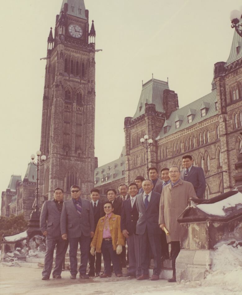 Thirteen men stand in a group on the steps in front of Canada's Parliament buildings.