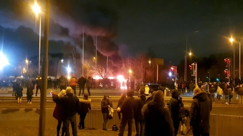 A group of people watch a fire outside a building in the distance.