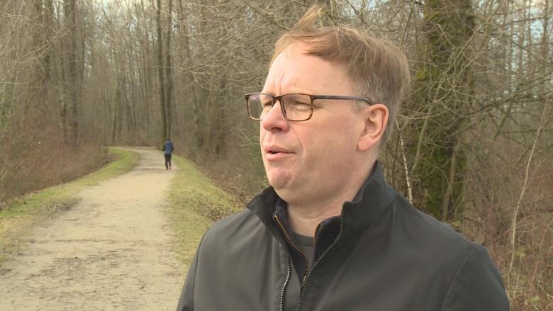 A white man in a black jacket speaks to a camera in a park.