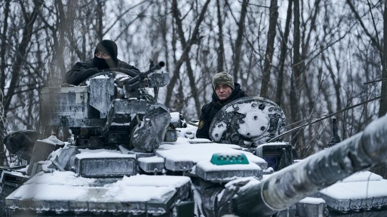 Two people are visible on top of a tank amidst trees. There is a light layer of snow on the tank.