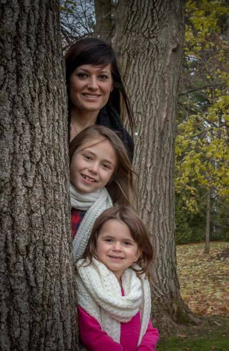 A mother stands outside with her two girls, all three of them poke their heads out from behind a tree. 