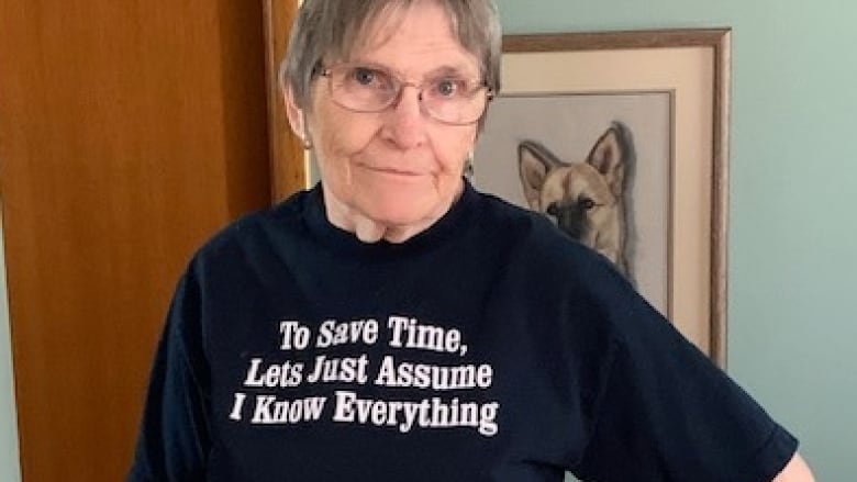 A woman in a black t-shirt and glasses looks into the camera. Her shirt reads: 