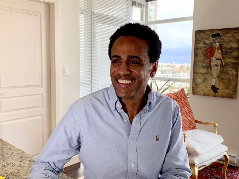 A man smiles in an apartment with a window behind him that looks out onto a city skyline and a framed portrait over his left shoulder.