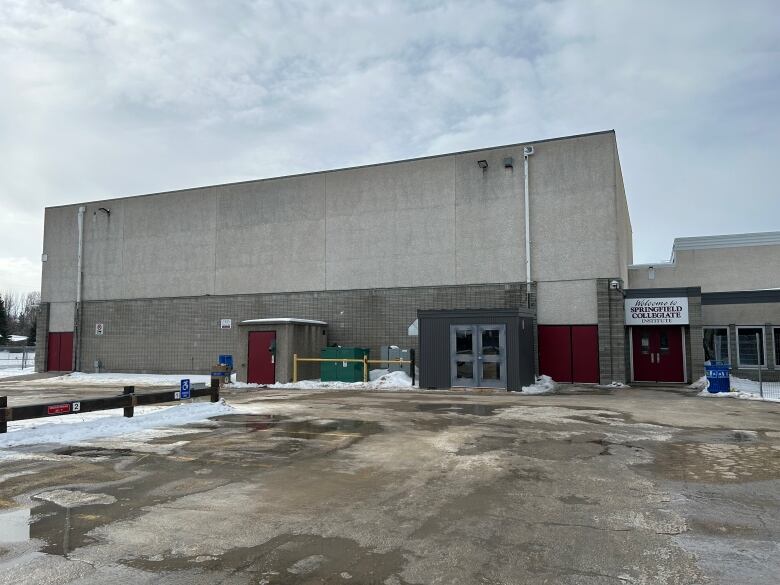 The grey cement and brick exterior of a high school gymnasium is pictured with a melting parking lot in the foreground.