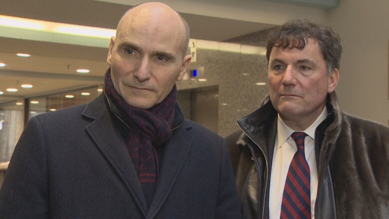 A bald man in a scarf and winter coat stands next to a man with short brown hair who is waiting a shirt and tie.