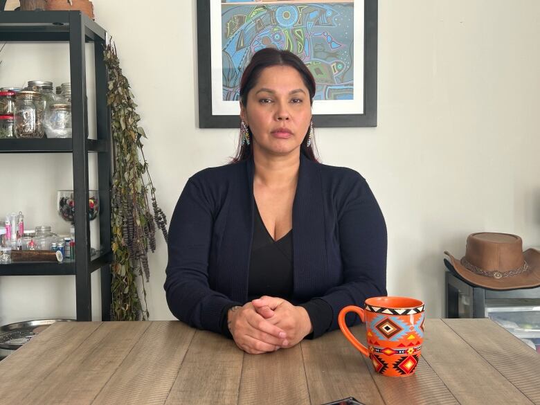 A woman in a blue sweater and shirt is pictured sitting down, holding her hands together at a wooden kitchen table. She has an orange coffee mug with First Nations artwork on the mug and in the background. She's sitting next to a shelf which contains traditional Indigenous medicines.   