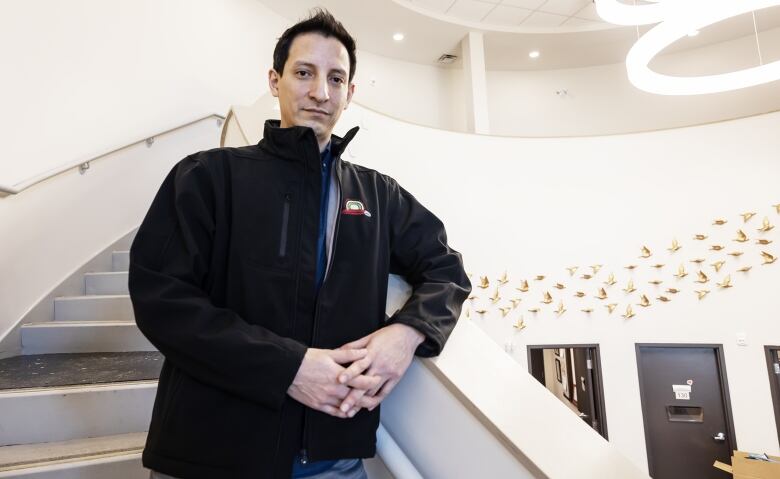 A man in a black jacket holds his hands together while standing on a curved staircase in a white foyer with gold decorative birds pictured on the wall in the background.